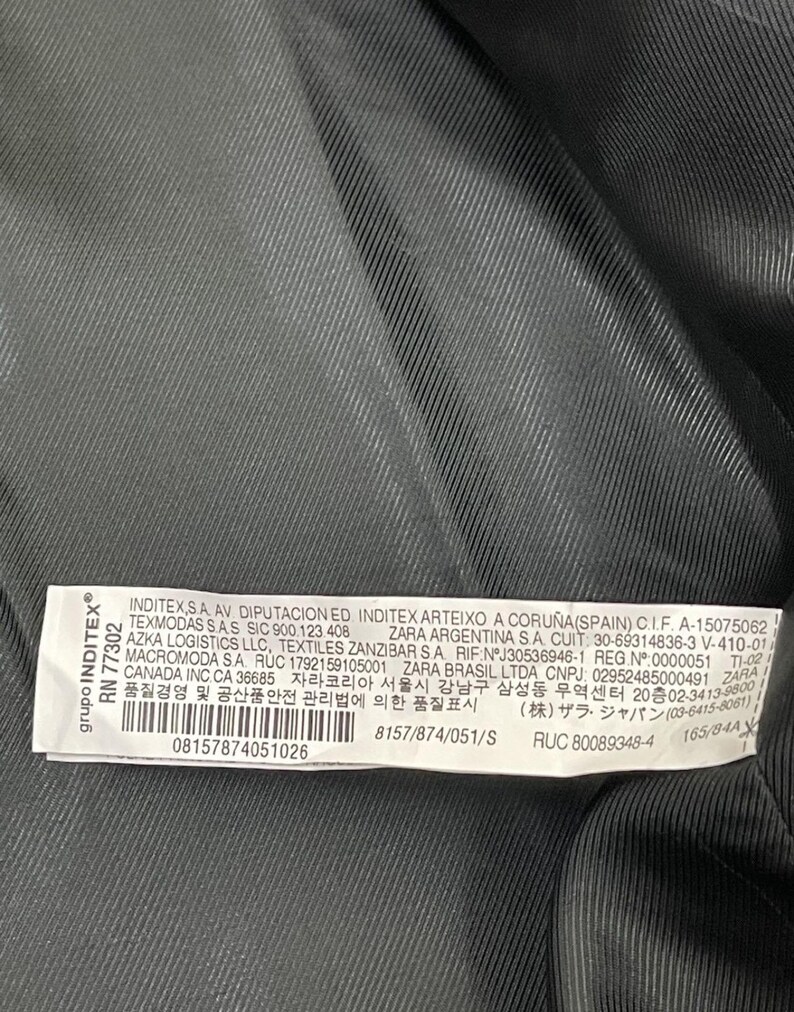 May include: Close-up of a garment label with text. The label is white with black text and is attached to a dark gray fabric. The label includes company information and product details.