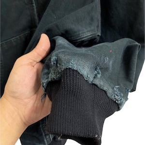 May include: Close-up of a black jacket sleeve with a ribbed cuff. The jacket fabric shows signs of wear and tear, with frayed edges. The sleeve is held up, showcasing the texture and details of the garment.