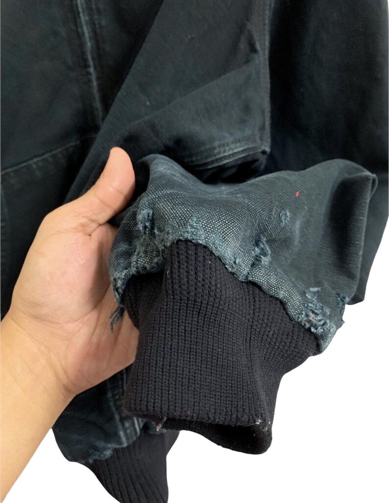 May include: Close-up of a black jacket sleeve with a ribbed cuff. The jacket fabric shows signs of wear and tear, with frayed edges. The sleeve is held up, showcasing the texture and details of the garment.