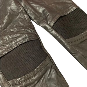 May include: Close-up of brown leather pants with ribbed knit panels at the knees. The leather has a slightly distressed appearance with visible stitching details. The pants appear to be a dark brown color.
