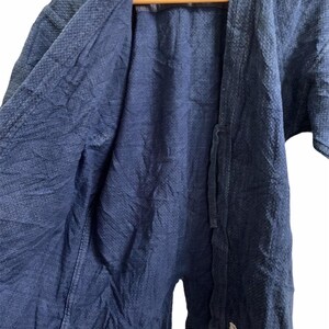 May include: A dark blue, open-front jacket with a textured weave. The jacket has short sleeves and a tie closure. The interior lining is a darker shade of blue. The garment is hanging on a wooden hanger.