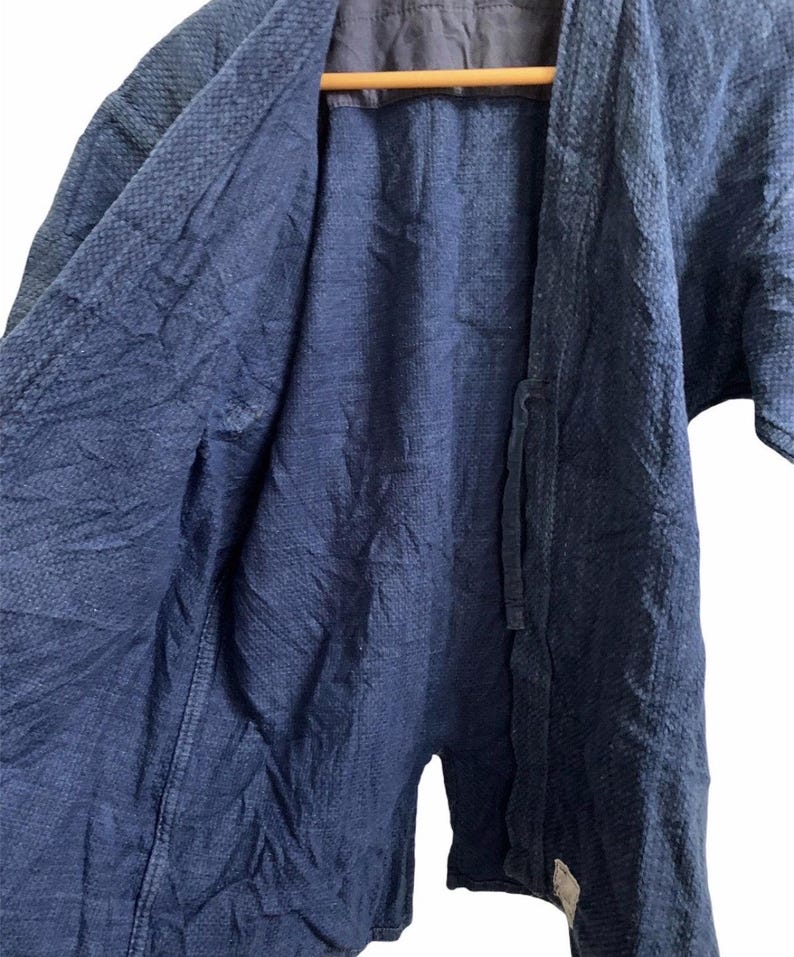 May include: A dark blue, open-front jacket with a textured weave. The jacket has short sleeves and a tie closure. The interior lining is a darker shade of blue. The garment is hanging on a wooden hanger.