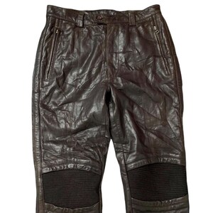 May include: Dark brown leather pants with a classic design. The pants feature two front pockets with zippers, a button closure, and a textured panel on the knees. The leather has a slightly wrinkled appearance.