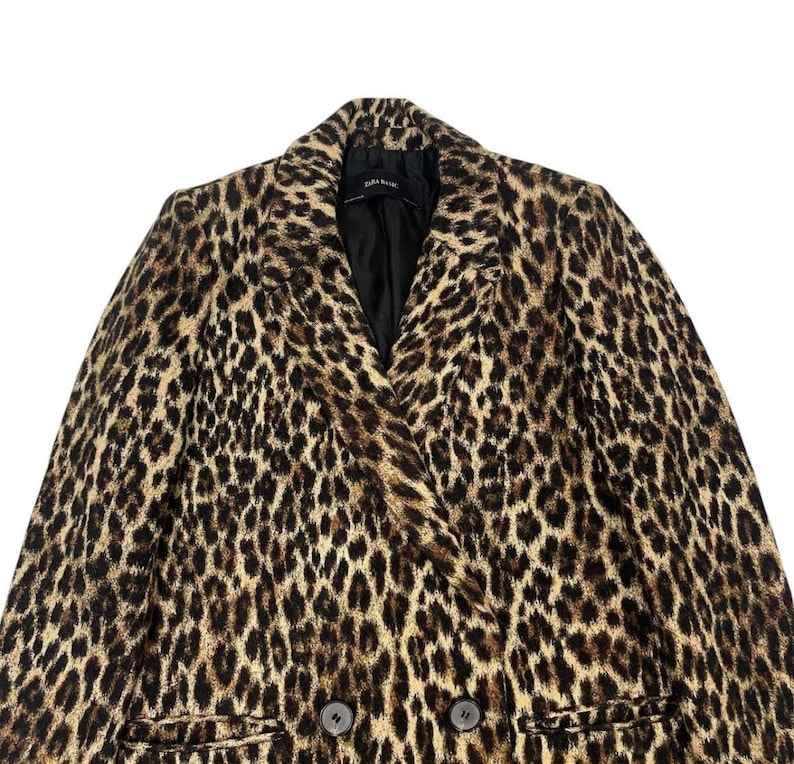 May include: A double-breasted leopard print coat with a notched collar and two dark buttons. The coat features a black lining and a classic, timeless design. The coat is a neutral color with brown and black spots.