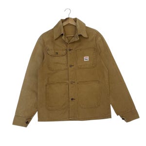 Puede incluir: Chaqueta de trabajo color canela con cierre de botones, dos bolsillos en el pecho y dos bolsillos inferiores. La chaqueta tiene cuello y un pequeño parche en el pecho derecho. La chaqueta está colgada en una percha de madera.