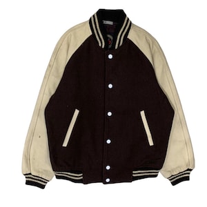 Könnte beinhalten: Eine Vintage-Collegejacke in Braun und Creme. Die Jacke hat cremefarbene Ärmel und einen braunen Körper mit weißen Akzenten. Sie hat einen schwarz-weiß gestreiften Kragen, Bündchen und Saum. Die Jacke hat weiße Knöpfe und zwei Vordertaschen.