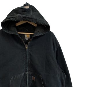 May include: Black hooded zip-up jacket with a distressed hood. The jacket features a front pocket and a brown logo patch. The jacket is hanging on a wooden hanger. The jacket is made of a durable material.