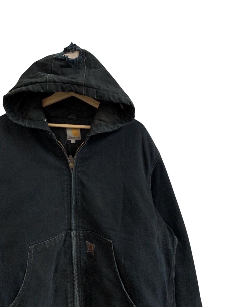 May include: Black hooded zip-up jacket with a distressed hood. The jacket features a front pocket and a brown logo patch. The jacket is hanging on a wooden hanger. The jacket is made of a durable material.