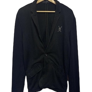 May include: Black blazer in a textured knit fabric. The jacket features a single-button closure, notched lapels, and two front pockets. A small skull and crossbones emblem is on the left chest. The blazer is hanging on a wooden hanger.