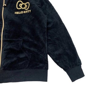 Puede incluir: Sudadera negra con cremallera dorada y el logotipo de Hello Kitty bordado en el pecho. La sudadera con capucha tiene una textura suave y aterciopelada, una capucha y un bolsillo delantero. Los pu&ntilde;os y el dobladillo son acanalados.