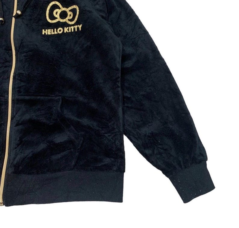 Puede incluir: Sudadera negra con cremallera dorada y el logotipo de Hello Kitty bordado en el pecho. La sudadera con capucha tiene una textura suave y aterciopelada, una capucha y un bolsillo delantero. Los pu&ntilde;os y el dobladillo son acanalados.