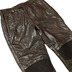 May include: Brown leather pants with zippered pockets and ribbed knee panels. The pants have a button and zipper closure at the waist. The leather has a slightly distressed appearance.