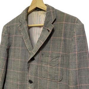 Op de afbeelding: Een grijze geruite blazer met een roze rasterpatroon. De blazer heeft een revers, twee voorzakken en een sluiting met twee knopen. De binnenvoering heeft verticale strepen in rood, wit en blauw.