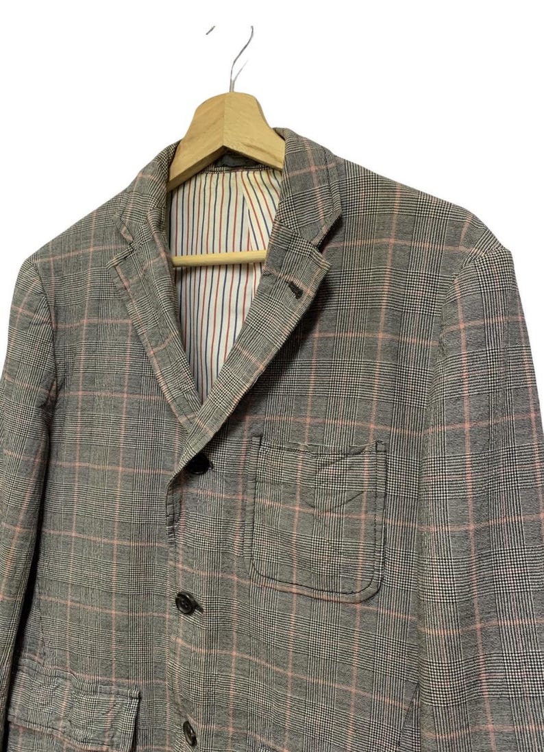 Op de afbeelding: Een grijze geruite blazer met een roze rasterpatroon. De blazer heeft een revers, twee voorzakken en een sluiting met twee knopen. De binnenvoering heeft verticale strepen in rood, wit en blauw.