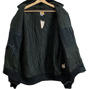 May include: Black Carhartt hooded jacket with a quilted interior, displayed open on a wooden hanger. The jacket has a ribbed hem and cuffs, with visible wear and tear on the fabric. The Carhartt logo is visible on a tag inside the jacket.