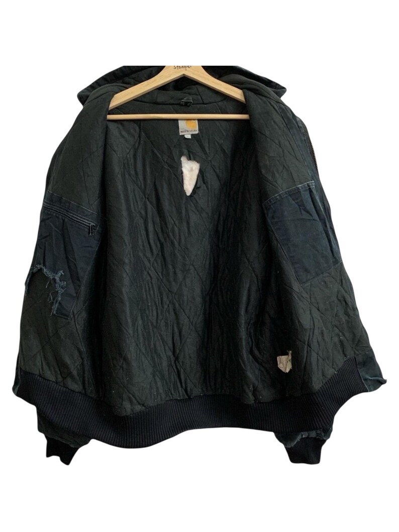 May include: Black Carhartt hooded jacket with a quilted interior, displayed open on a wooden hanger. The jacket has a ribbed hem and cuffs, with visible wear and tear on the fabric. The Carhartt logo is visible on a tag inside the jacket.