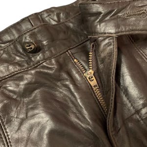 May include: Close-up of brown leather pants with a visible zipper and button closure. The leather appears worn and textured, with visible creases and folds. The zipper is a brass color, and the button is dark brown.