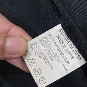 Puede incluir: Una etiqueta blanca con texto negro, sujeta contra una tela negra. La etiqueta dice "FABRIQUE AU JAPON MADE IN JAPAN 100% COTON COTTON" e incluye instrucciones de cuidado con s&iacute;mbolos.