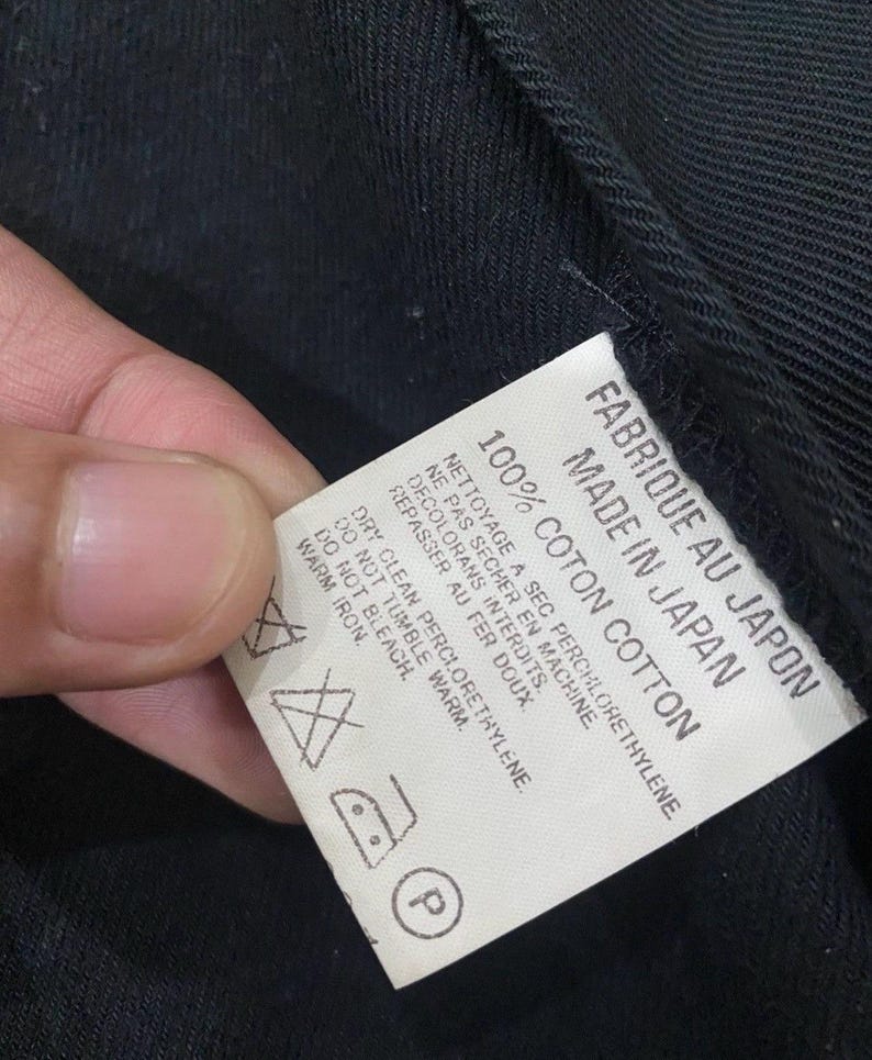 Puede incluir: Una etiqueta blanca con texto negro, sujeta contra una tela negra. La etiqueta dice "FABRIQUE AU JAPON MADE IN JAPAN 100% COTON COTTON" e incluye instrucciones de cuidado con s&iacute;mbolos.