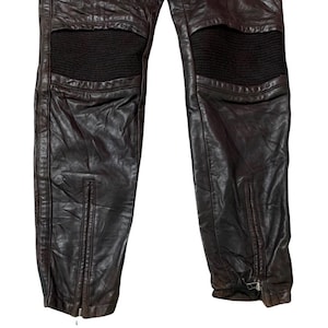 May include: Close-up of brown leather pants with black padded knee sections. The pants have zippers at the ankles and show signs of wear. The leather appears distressed, giving a vintage look.