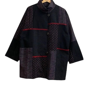Könnte beinhalten: Eine Patchwork-Jacke in Schwarz-, Marine- und Burgundertönen mit Stehkragen und Knopfverschluss. Das Design integriert verschiedene strukturierte Stoffe, darunter gepunktete und gestreifte Muster, mit dünnen roten Akzentlinien. Die Jacke wird auf einem Holzbügel präsentiert.