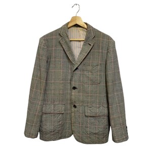 Op de afbeelding: Een grijze blazer met een roze ruitpatroon, drie zwarte knopen en twee voorzakken. De blazer hangt aan een houten hanger. De binnenvoering heeft een streepjespatroon.
