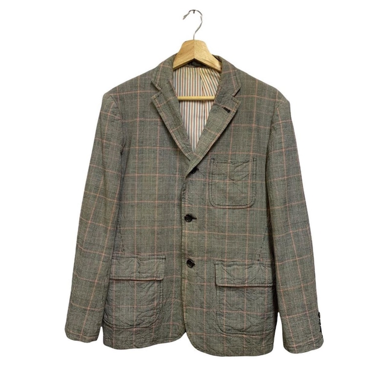 Op de afbeelding: Een grijze blazer met een roze ruitpatroon, drie zwarte knopen en twee voorzakken. De blazer hangt aan een houten hanger. De binnenvoering heeft een streepjespatroon.