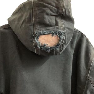 May include: Close-up of a dark gray hooded jacket with a distressed, worn appearance. The hood features a large hole at the back, revealing the inner lining. The jacket appears to be made of a durable fabric, with visible stitching.