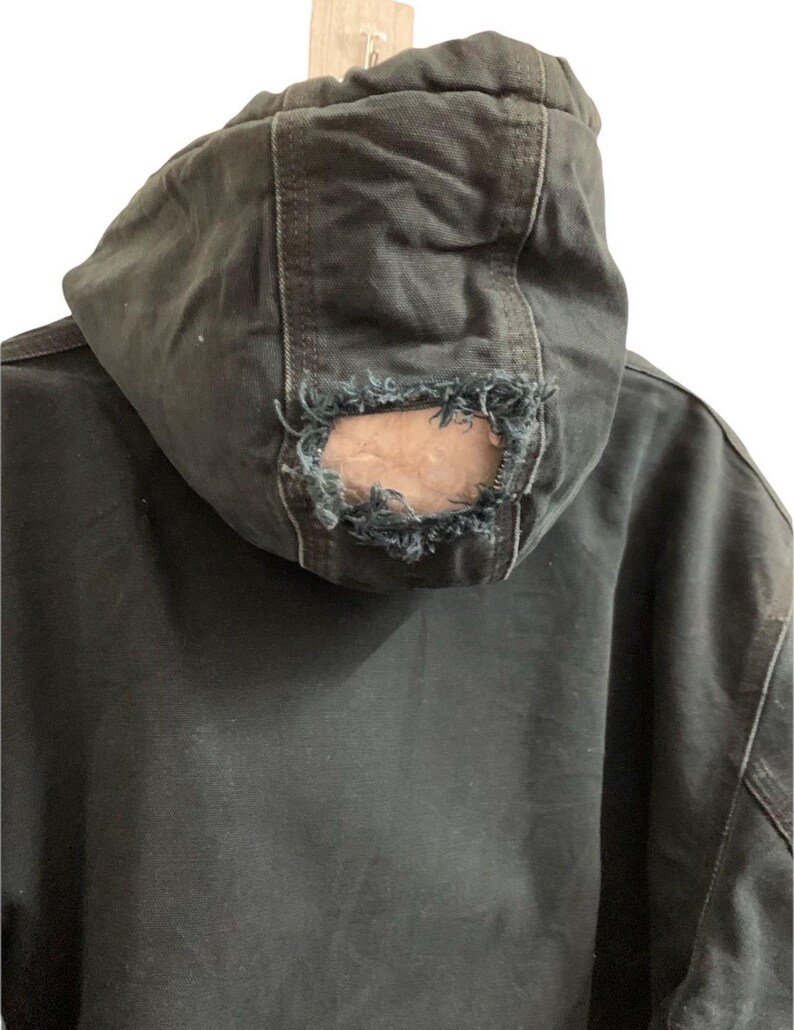May include: Close-up of a dark gray hooded jacket with a distressed, worn appearance. The hood features a large hole at the back, revealing the inner lining. The jacket appears to be made of a durable fabric, with visible stitching.