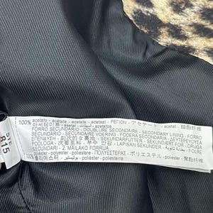 May include: Close-up of the inside of a coat. The lining is black, with a ribbed texture. A tag with text in multiple languages is visible, listing fabric composition and care instructions. The outer fabric has a leopard print.