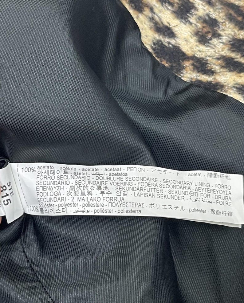 May include: Close-up of the inside of a coat. The lining is black, with a ribbed texture. A tag with text in multiple languages is visible, listing fabric composition and care instructions. The outer fabric has a leopard print.