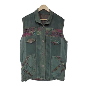 May include: A green corduroy vest with floral patchwork accents. The vest features a button-up front, two chest pockets, and two lower pockets. The floral patches include pink, purple, and green patterns. The vest is sleeveless and hangs on a wooden hanger.