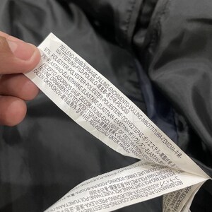 Puede incluir: Primer plano de una etiqueta de ropa blanca con texto negro. La etiqueta est&aacute; sujeta por una mano y est&aacute; contra una tela azul oscuro. La etiqueta incluye las palabras "FILLING", "POLYESTER" y "DECORATIONS".