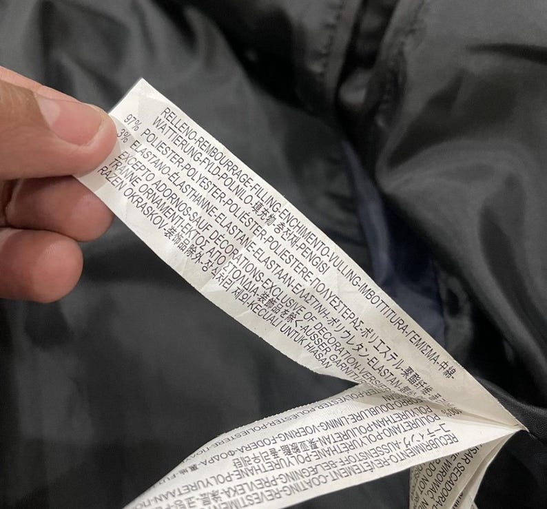 Puede incluir: Primer plano de una etiqueta de ropa blanca con texto negro. La etiqueta est&aacute; sujeta por una mano y est&aacute; contra una tela azul oscuro. La etiqueta incluye las palabras "FILLING", "POLYESTER" y "DECORATIONS".