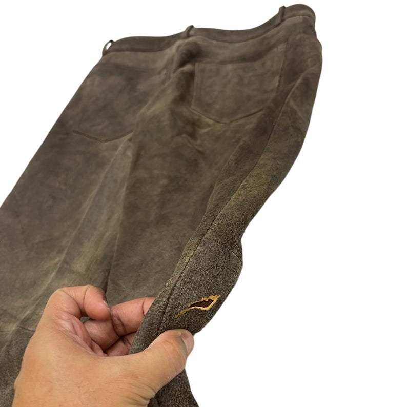 Pode incluir: Close-up de cal&ccedil;as de camur&ccedil;a castanhas com dois bolsos traseiros. As cal&ccedil;as t&ecirc;m um aspeto gasto e envelhecido. Uma m&atilde;o segura a pe&ccedil;a, mostrando a textura e as varia&ccedil;&otilde;es de cor da camur&ccedil;a.