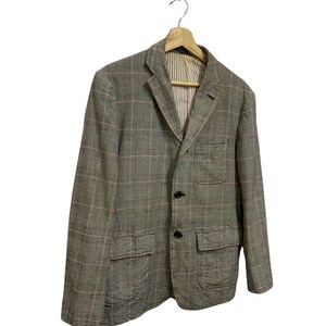 Op de afbeelding: Een grijze blazer met een roze en wit ruitpatroon, hangend aan een houten hanger. De jas heeft twee voorzakken en een gestreepte voering. Hij is voorzien van zwarte knopen.