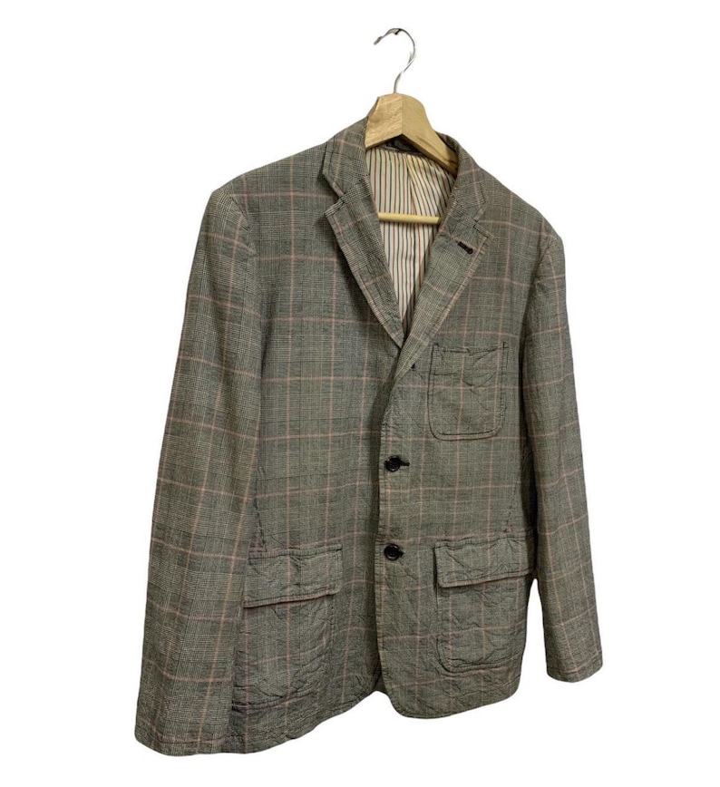 Op de afbeelding: Een grijze blazer met een roze en wit ruitpatroon, hangend aan een houten hanger. De jas heeft twee voorzakken en een gestreepte voering. Hij is voorzien van zwarte knopen.
