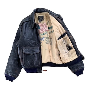 May include: A vintage black leather bomber jacket with a cream lining. The jacket has two front pockets, a zip closure, and a navy blue ribbed collar and cuffs. The lining features a graphic print with text and an American flag.