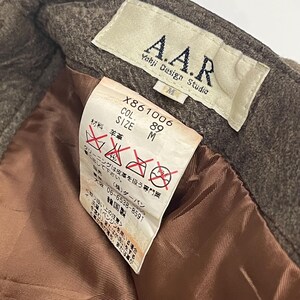 Pode incluir: Close-up de uma etiqueta de vestu&aacute;rio com o texto "A.A.R Yohji Design Studio". A etiqueta inclui instru&ccedil;&otilde;es de cuidado e informa&ccedil;&otilde;es de tamanho. A pe&ccedil;a de vestu&aacute;rio parece ser feita de l&atilde;. A etiqueta est&aacute; presa a um tecido castanho.