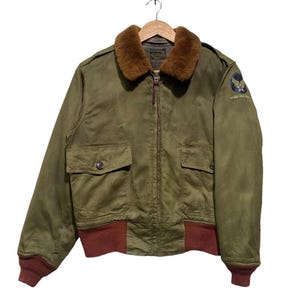 May include: Olive green bomber jacket with a brown faux fur collar, two front pockets, and a brown ribbed hem and cuffs. Features a patch on the upper left sleeve with a star and text. The jacket is hanging on a wooden hanger.