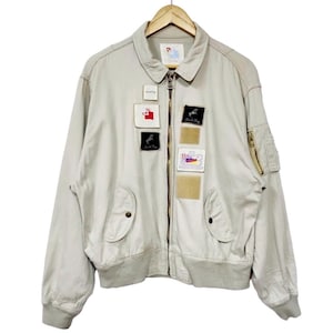 May include: A beige bomber jacket with a zip-up front, several patches, and a ribbed hem and cuffs. The jacket is suspended on a wooden hanger. The patches display diverse designs and text.