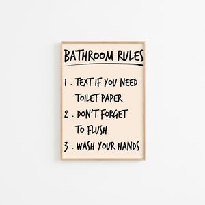 Puede incluir: Impresión enmarcada con el texto "BATHROOM RULES" en negro. Las reglas son: "1. TEXT IF YOU NEED TOILET PAPER", "2. DON'T FORGET TO FLUSH" y "3. WASH YOUR HANDS". El fondo es de color beige claro.