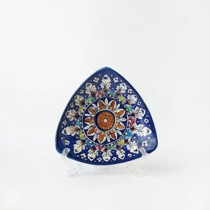 Może przedstawiać: Trójkątny, ceramiczny talerz z kwiatowym wzorem. Talerz ma granatowe tło z centralnym pomarańczowo-brązowym kwiatem otoczonym kolorowymi płatkami i białymi akcentami. Talerz jest prezentowany na przezroczystym stojaku.