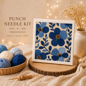 Op de afbeelding: Een punch needle borduurpakket met een bloemenontwerp van blauwe en marineblauwe bloemen. De kit bevat bollen garen in verschillende blauwtinten, een punch needle tool en de tekst "PUNCH NEEDLE KIT EMBROIDERY KIT CREATE • RELAX • ENJOY."