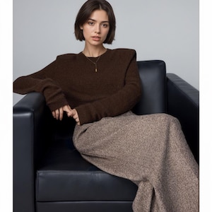 May include: A dark brown long-sleeved knit sweater with structured shoulders and a crew neckline, paired with a long, textured beige and brown speckled skirt. A thin gold-toned necklace with a small rectangular pendant is worn.