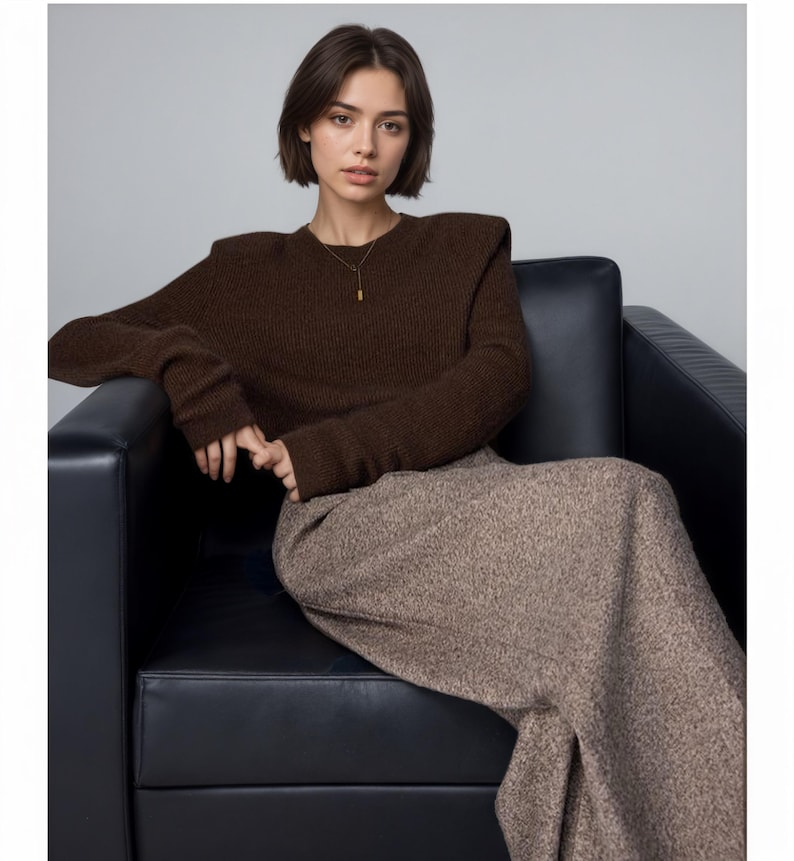 May include: A dark brown long-sleeved knit sweater with structured shoulders and a crew neckline, paired with a long, textured beige and brown speckled skirt. A thin gold-toned necklace with a small rectangular pendant is worn.