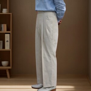 Op de afbeelding: Lichtbeige geplooide broek met een relaxte pasvorm. De broek wordt gecombineerd met lichtgrijze loafers. Het model draagt een lichtblauw overhemd met knopen. De achtergrond is neutraal bruin.