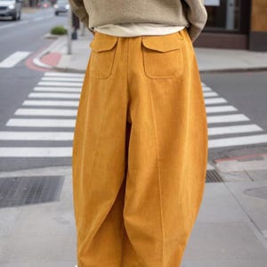 May include: Mustard yellow corduroy trousers featuring a wide-leg silhouette, two back patch pockets with flaps, and cuffed hems. The pants are paired with a beige long-sleeve top and white sneakers, showcasing a relaxed fit and textured fabric.