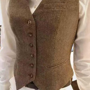 May include: A brown herringbone patterned vest featuring a V-neckline, five front buttons, and two welt pockets. The vest is worn over a crisp white collared shirt, showcasing a structured fit with a pointed hemline at the waist.