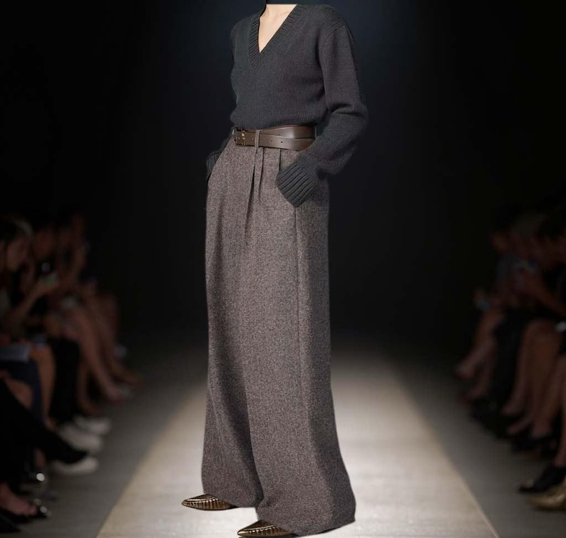 May include: A dark charcoal gray V-neck long-sleeve sweater paired with high-waisted, wide-leg brown tweed trousers. The trousers feature a brown leather belt with a buckle at the waist and are worn with pointed-toe metallic shoes.